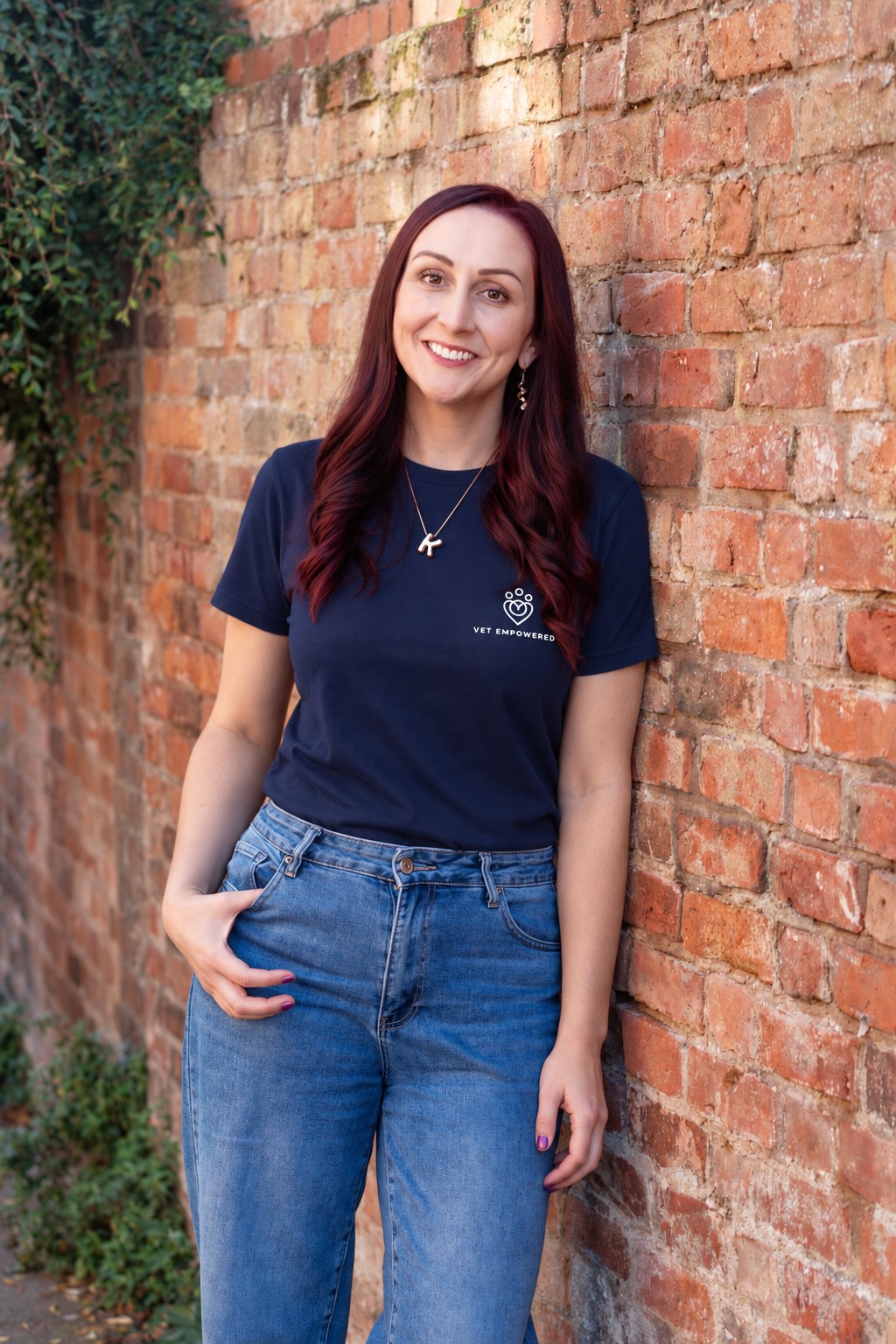 Dr Katie Ford smiling in a navy Vet Empowered T-shirt and jeans, leaning against a brick wall with climbing plants