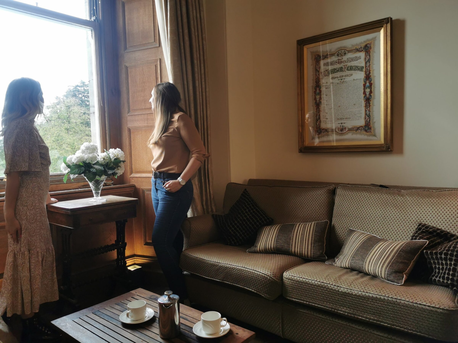 Katie and Claire looking out of a tall window, with coffee on the table