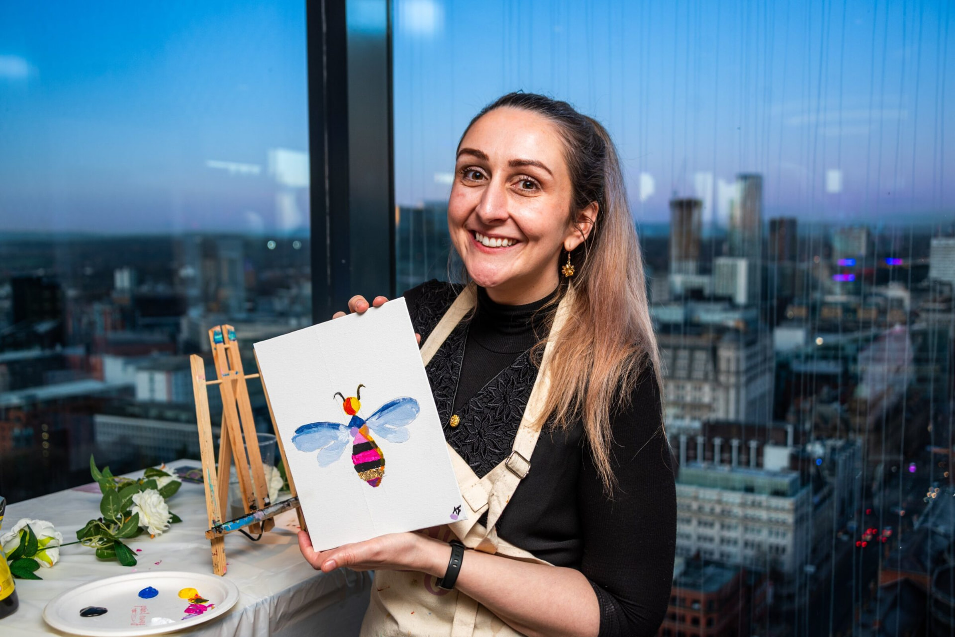 Katie holding up her painting of a bee, with the Manchester skyline behind